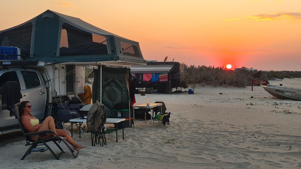 A women on a camp chair reclining in front of a tent at the beach with a sunset